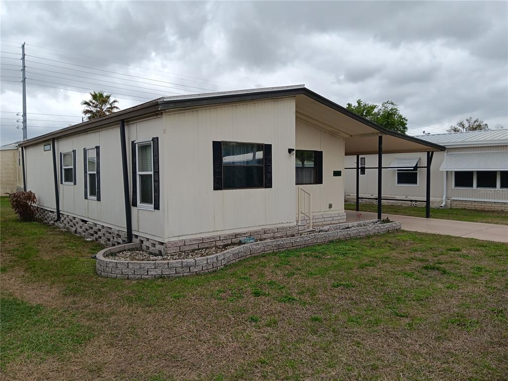A single-story mobile home with a carport, tan siding, and a low brick retaining wall — 37536 AMIGO DRIVE, ZEPHYRHILLS, FL