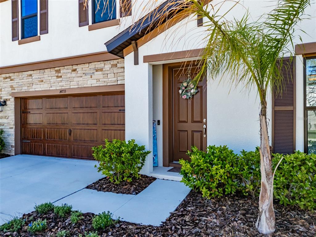 A white house exterior features a brown garage door, a front door with a wreath, and landscaping — 14733 GALLOP RUN DRIVE, LITHIA, FL