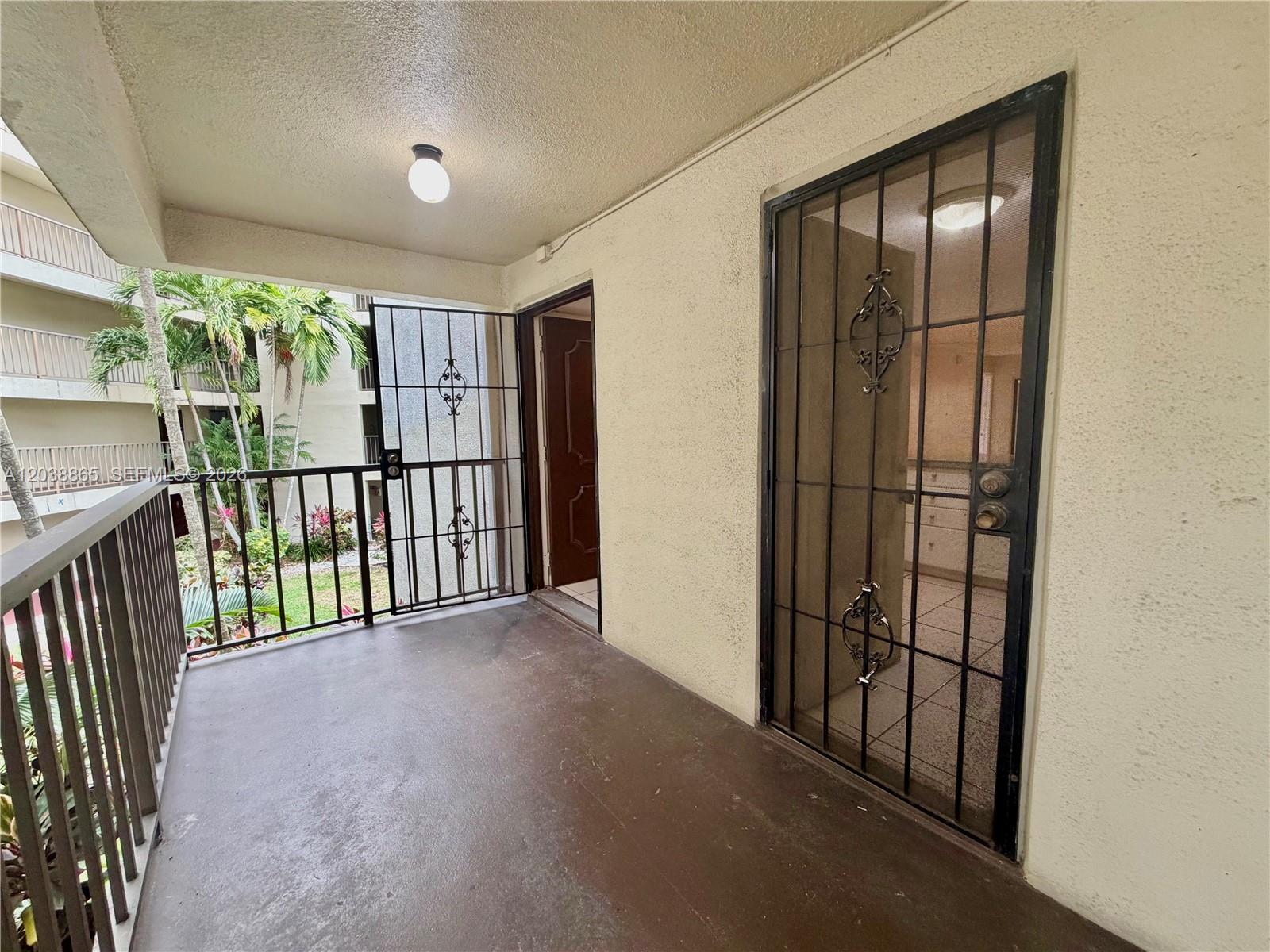 A balcony features a metal security door, a brown floor, and a view of palm trees — 2320 S Cypress Bend Dr # 206, Pompano Beach FL 33069, POMPANO BEACH, FL