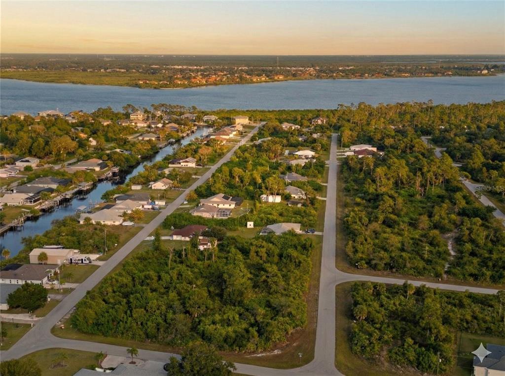 An aerial view shows residential houses, a canal, and undeveloped wooded lots near a bay — 12482 KNEELAND TERRACE, PORT CHARLOTTE, FL