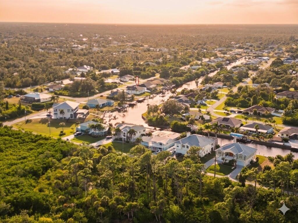 An aerial view shows a waterfront neighborhood with houses, canals, and surrounding trees at sunset — 12482 KNEELAND TERRACE, PORT CHARLOTTE, FL