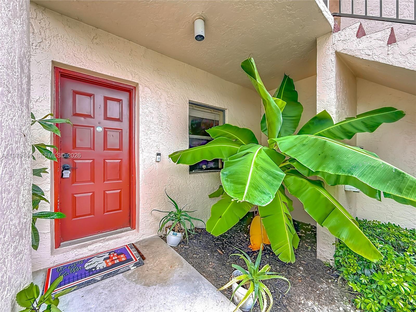 A red front door sits next to a large banana plant on a concrete porch — 8422 E Boca Glades Blvd E # 8422, Boca Raton FL 33434, BOCA RATON, FL