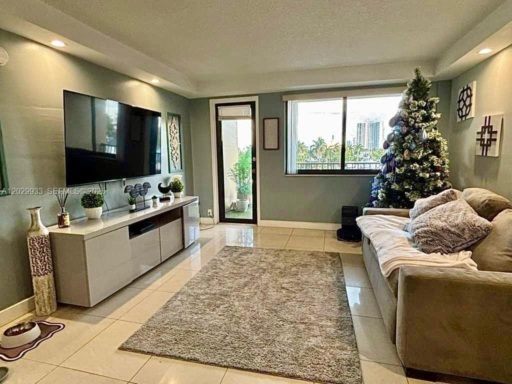 A living room features a large television, a gray sofa, and a Christmas tree — 301 Golden Isles Dr # 406, Hallandale Beach FL 33009, HALLANDALE BEACH, FL