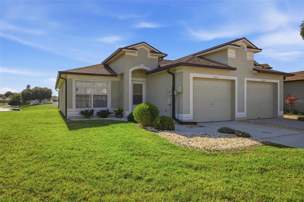A tan, single-story duplex with two garage doors and a green lawn under a blue sky — 13307 ASHBARK COURT, RIVERVIEW, FL