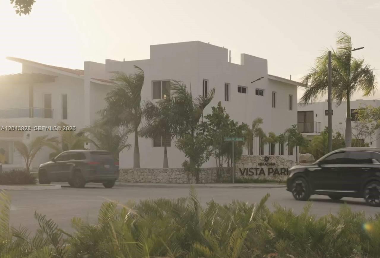 A white multi-story building stands behind a stone wall with palm trees and two cars — vistta cana, FL