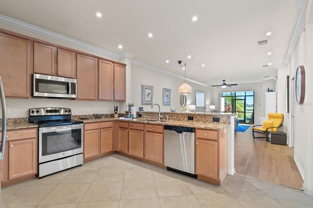 A kitchen with light wood cabinets, granite countertops, and stainless steel appliances overlooks a living area — 131 BABBLING BROOK RUN, BRADENTON, FL