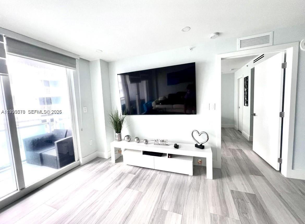 A modern living room features a wall-mounted television, white media console, and light wood flooring — 6917 Collins Ave # 806, Miami Beach FL 33141, MIAMI BEACH, FL