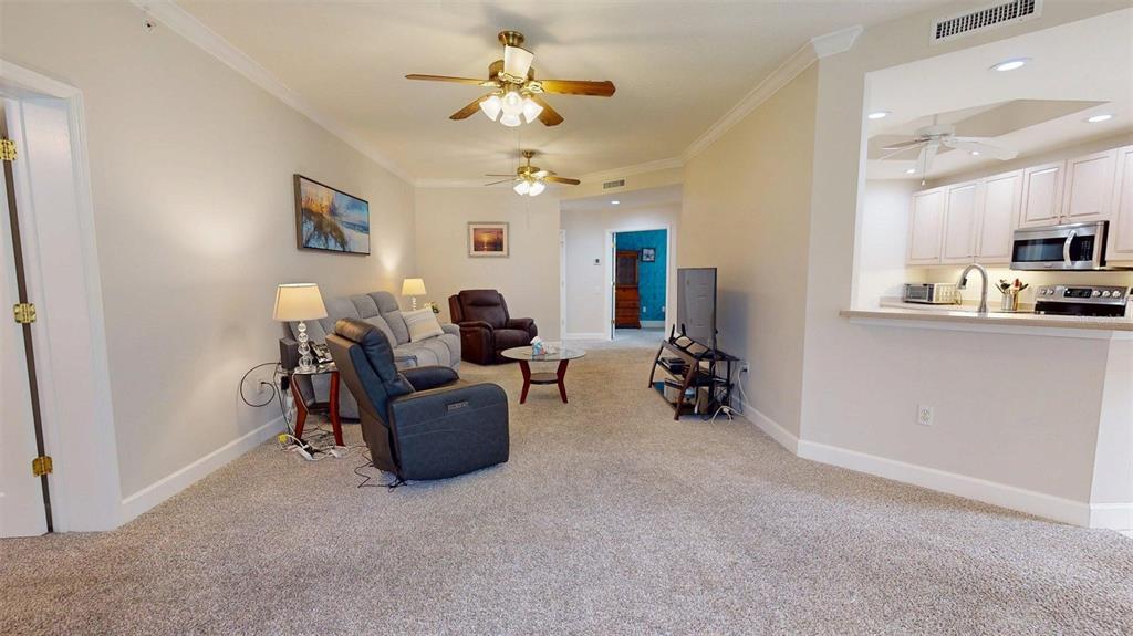 A living room with carpeted floors, two ceiling fans, and an open kitchen with white cabinets — 7434 BOTANICA PARKWAY UNIT 101, SARASOTA, FL