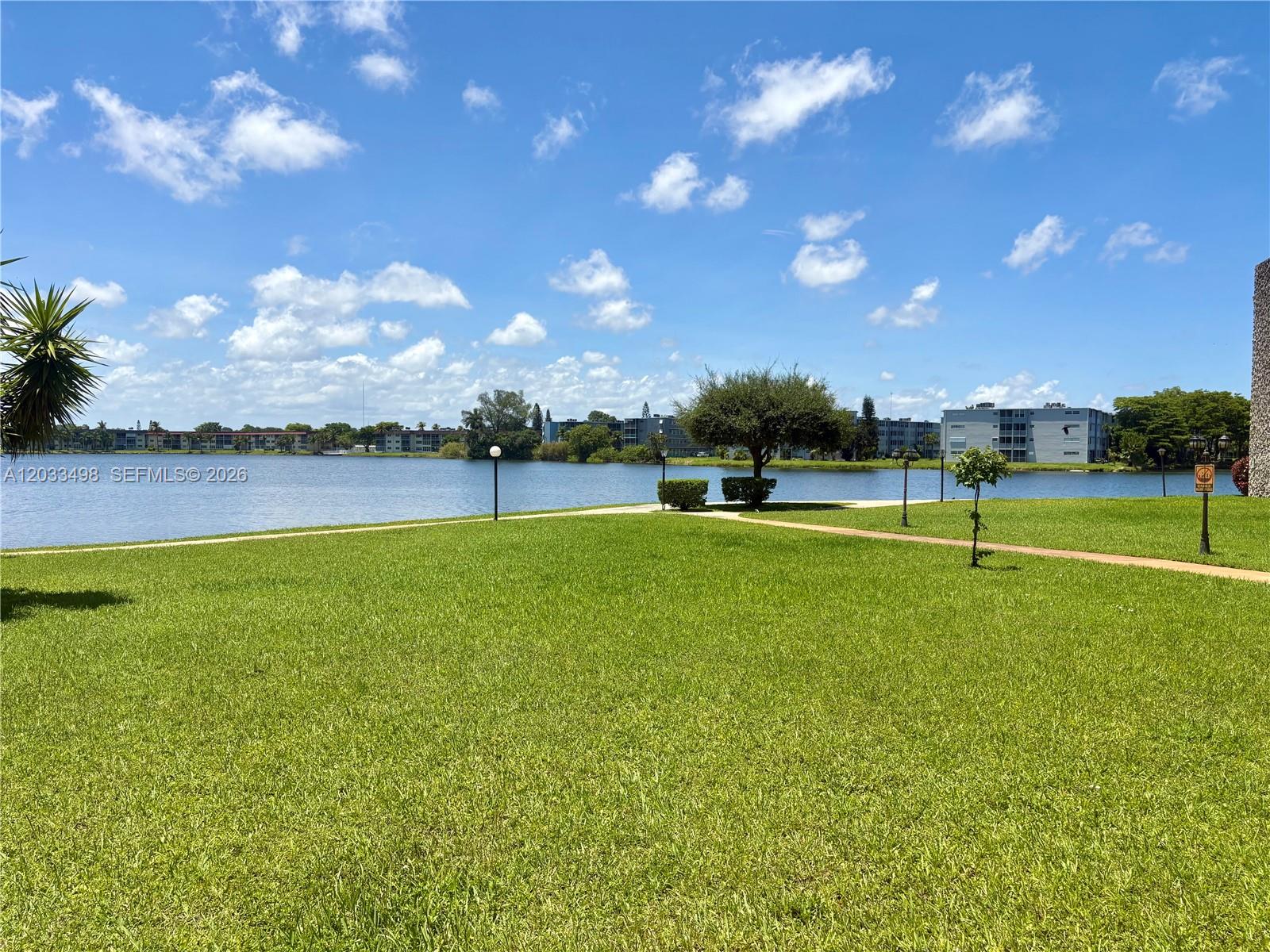 A grassy lawn overlooks a lake with apartment buildings in the background under a sky — MIAMI, FL