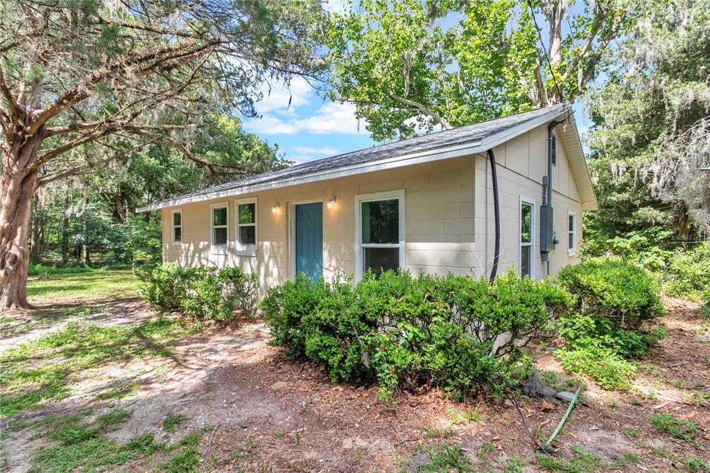 A small, light-colored concrete block house sits in a wooded yard with green bushes — 658 NE 39TH COURT, OCALA, FL