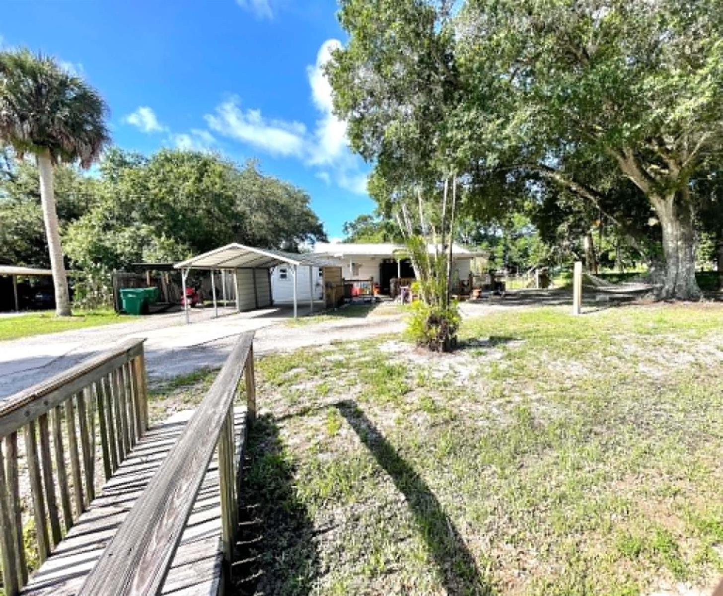 A wooden footbridge leads toward a house with a metal carport and large shade trees — 1111 NW 3rd Street, Okeechobee FL 34972, OKEECHOBEE, FL