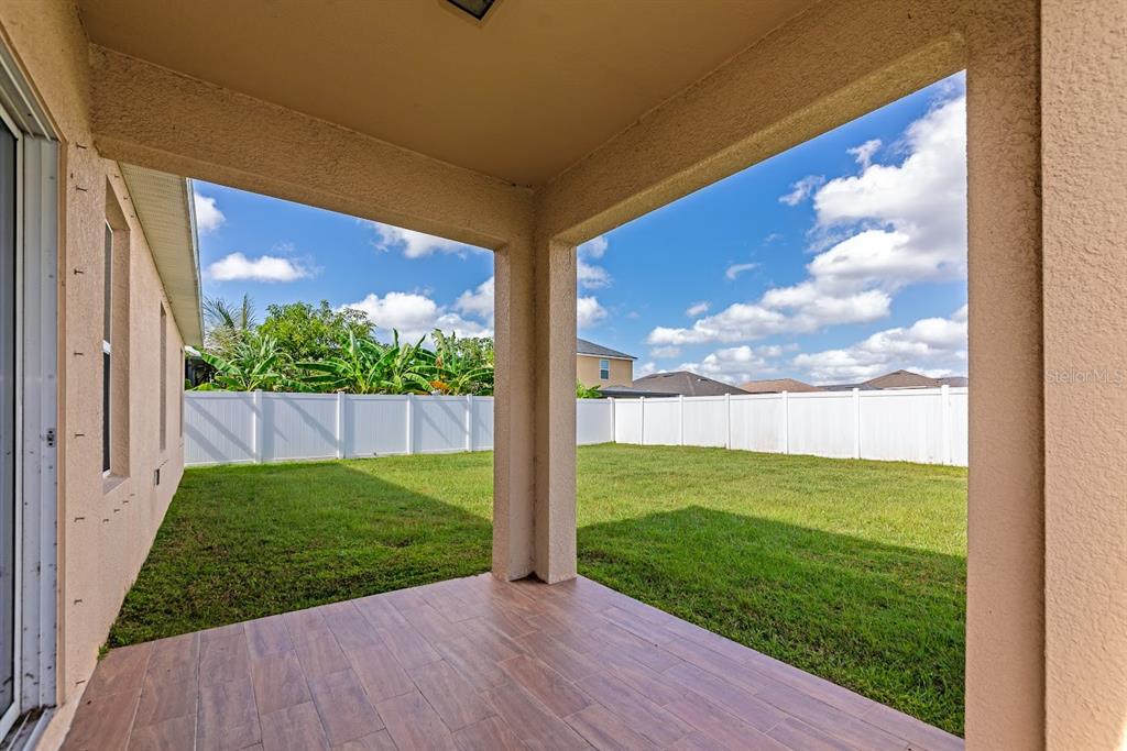 A covered patio with wood-look tile flooring overlooks a grassy backyard enclosed by a fence — 15517 ROSE GROVE DRIVE, BRADENTON, FL