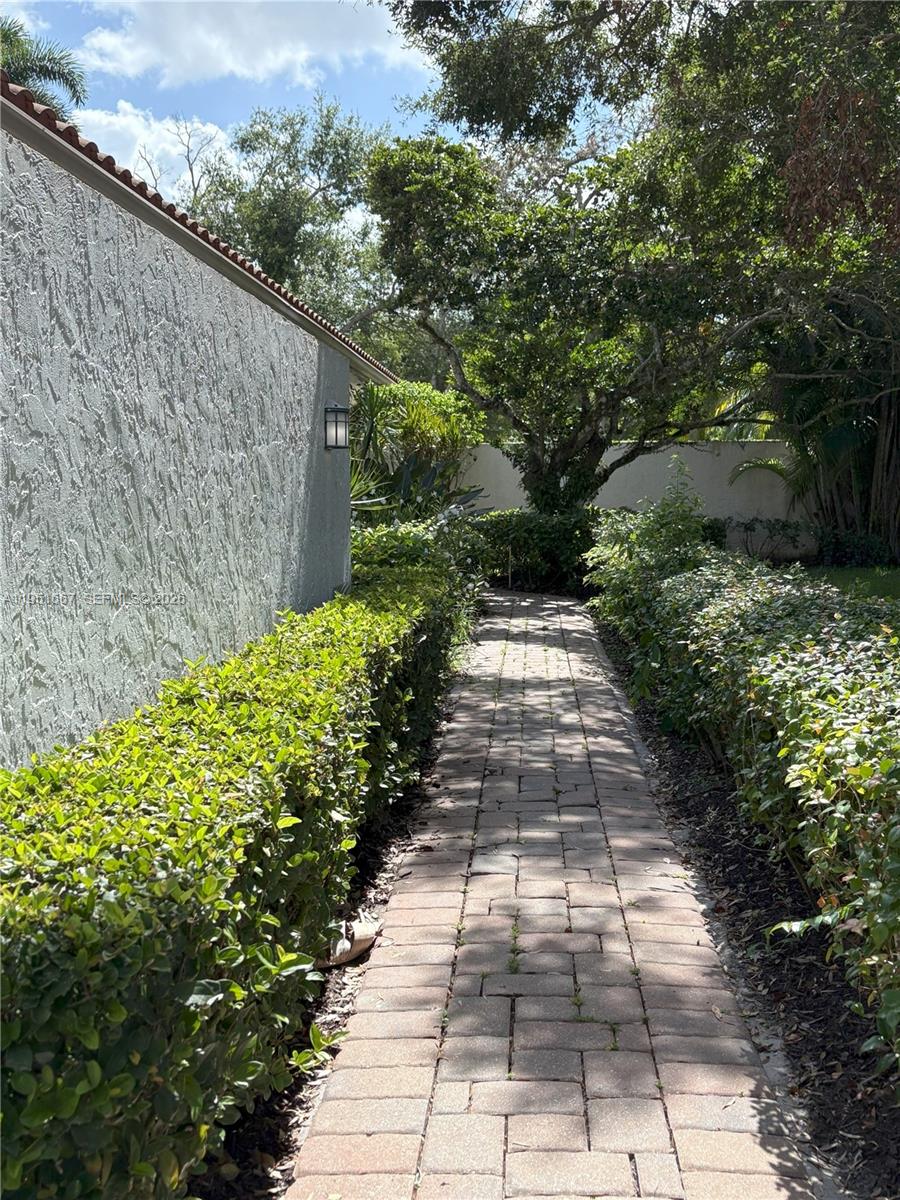 A paved walkway runs alongside a stucco wall between two rows of green hedges — 2538 Windsor Way Ct, Wellington FL 33414, WELLINGTON, FL