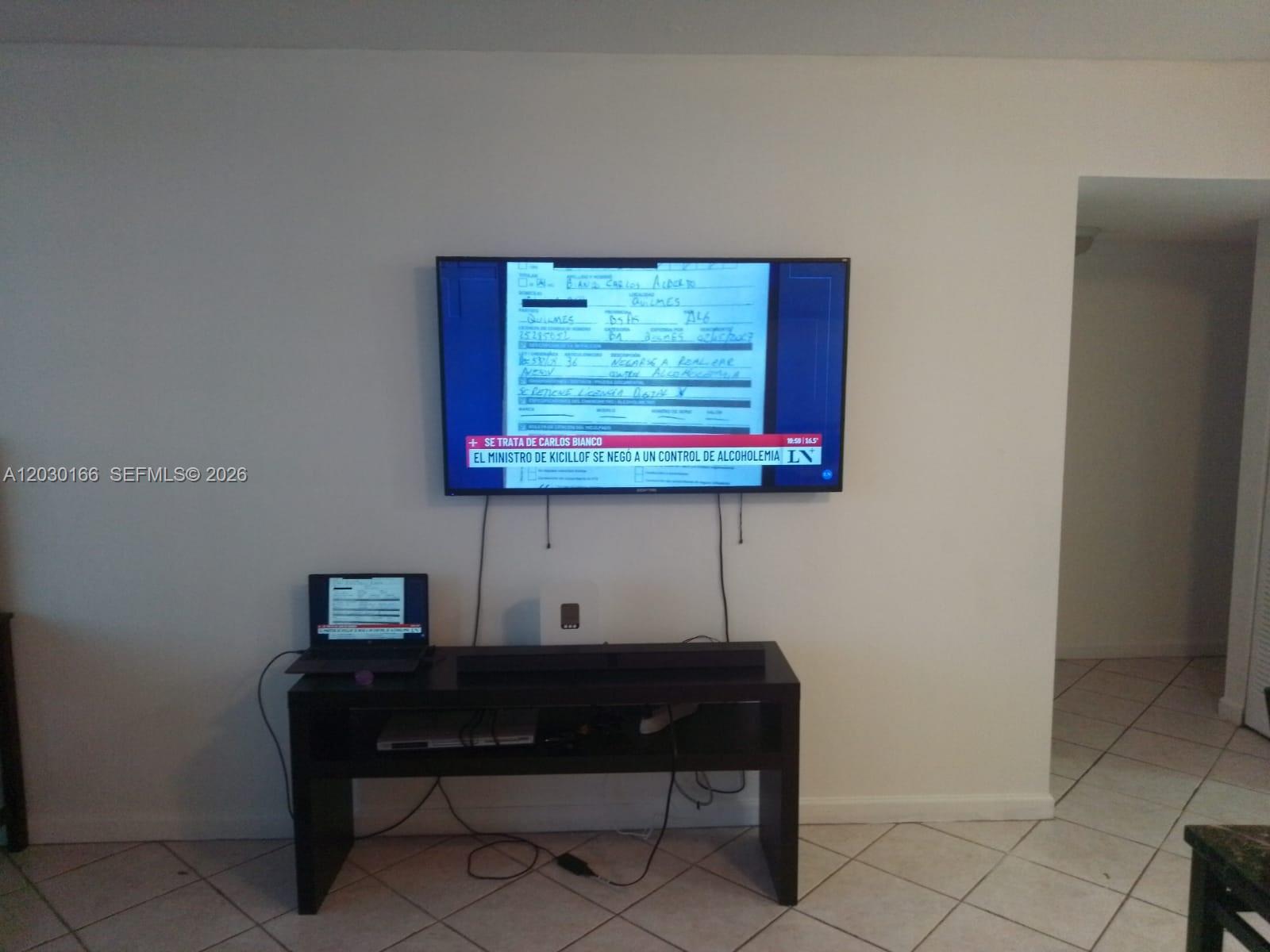 A wall-mounted television displays a news report above a black media console and laptop — 13390 NE 7th Ave # 207, North Miami FL 33161, NORTH MIAMI, FL