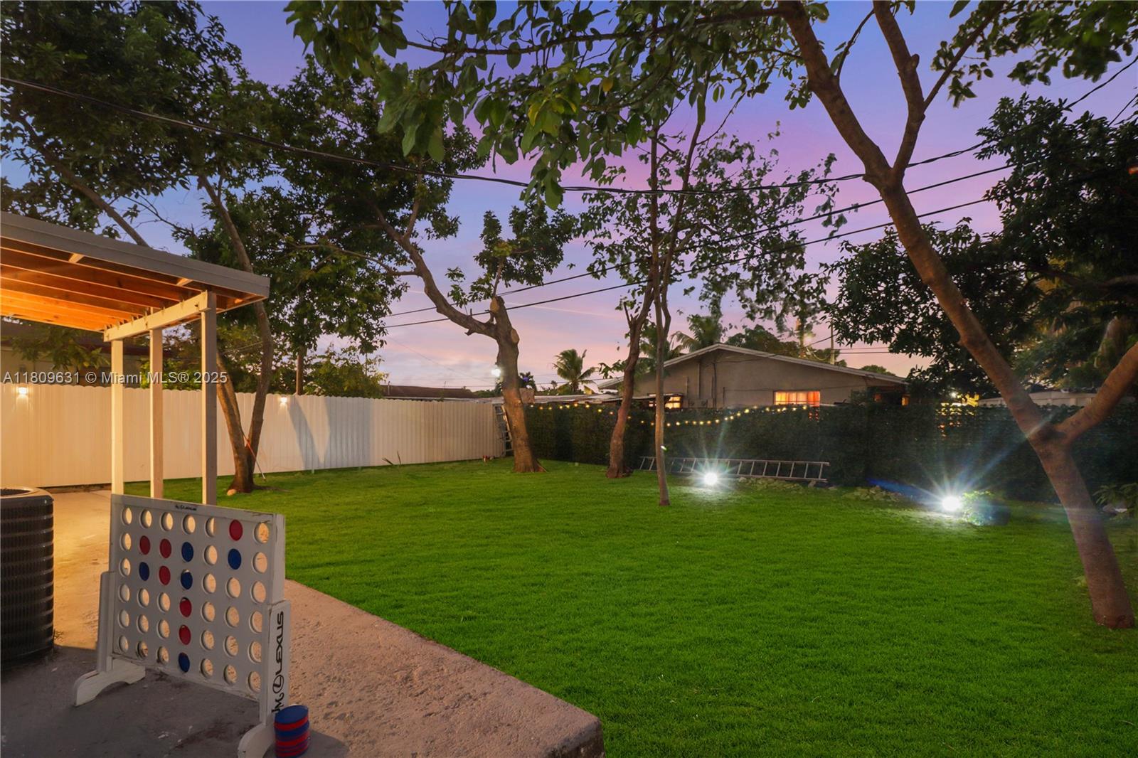 A backyard features a green lawn, mature trees, string lights, and a large game board — 6039 SW 38th St, Miramar FL 33023, MIRAMAR, FL
