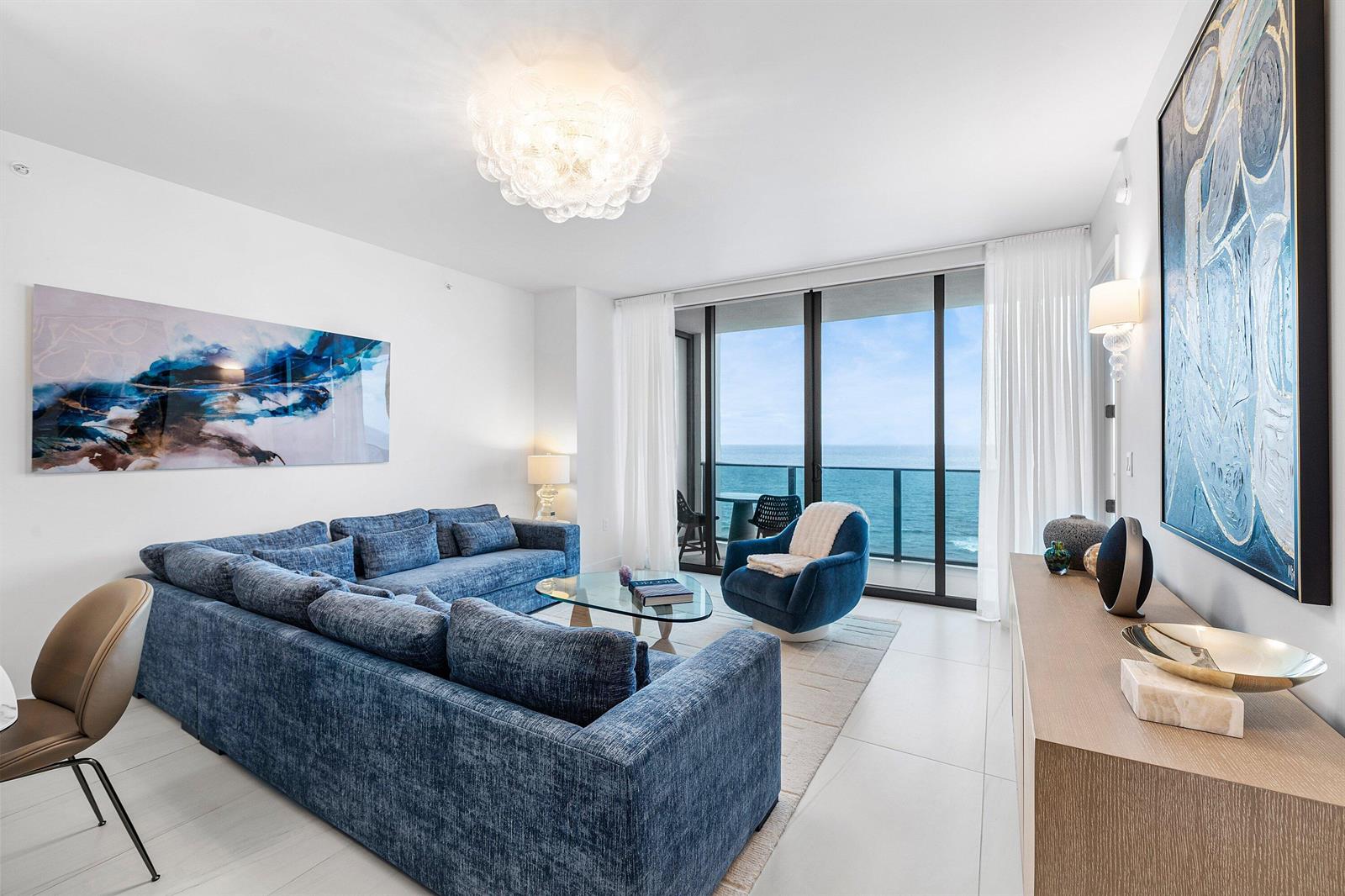 A living room features a blue sectional sofa, a glass coffee table, and ocean views — 900 N Ocean Boulevard # 1504, Pompano Beach FL 33062, POMPANO BEACH, FL