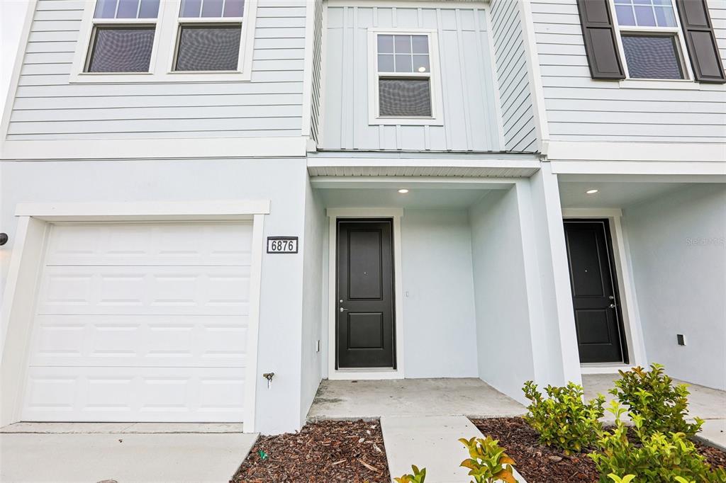 A two-story townhouse exterior features a white garage, a black front door, and light siding — 6876 HOLCOMBE DOWN DRIVE, ZEPHYRHILLS, FL
