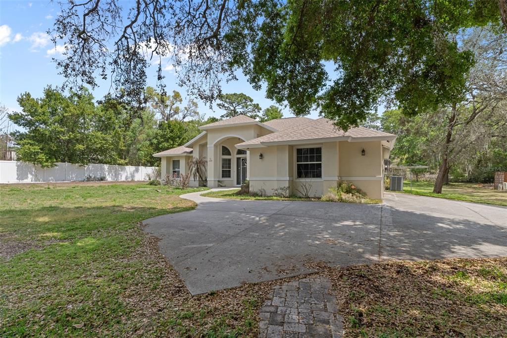 A single-story beige house with a concrete driveway and a brick walkway under large trees — 17948 GREENSBORO STREET, SPRING HILL, FL