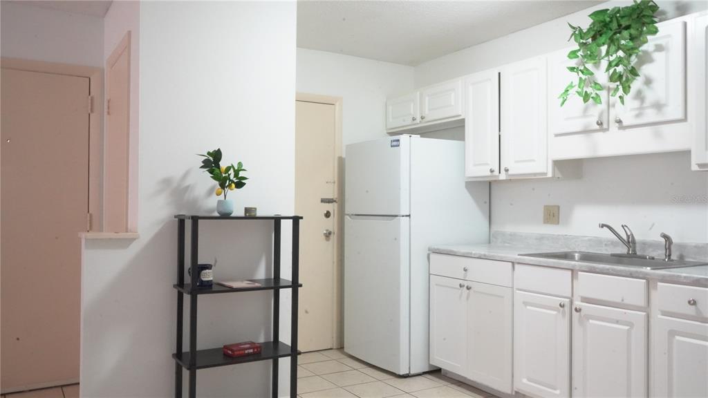 A small kitchen features white cabinets, a white refrigerator, and a black metal shelving unit — 1700 SW 16TH COURT UNIT J-4, GAINESVILLE, FL