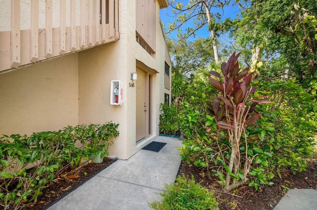 A beige exterior building entrance features a concrete walkway, a door, and lush landscaping — 5146 HARPERS CROFT UNIT 13, SARASOTA, FL