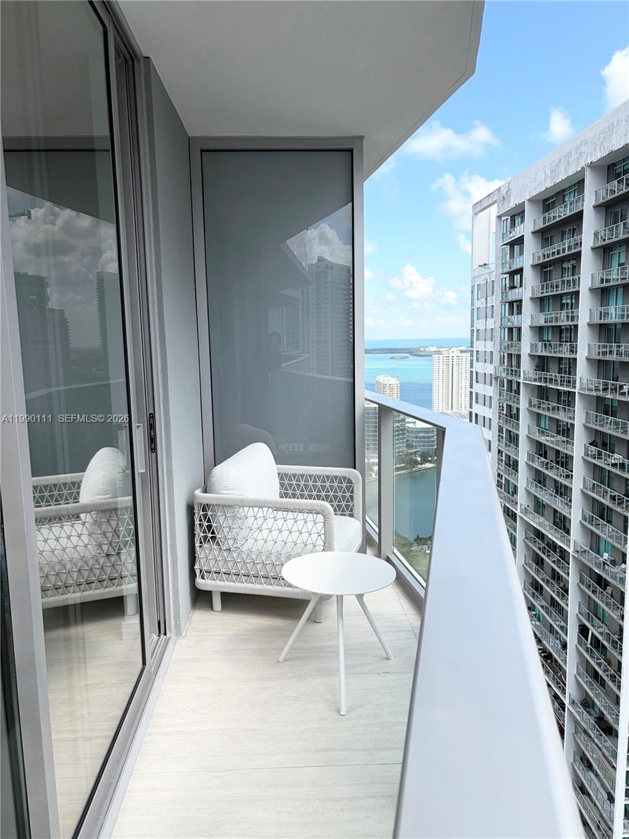 A high-rise balcony features a white woven chair, a small table, and glass railings — 77 SE 5th street # 2709, Miami FL 33131, MIAMI, FL
