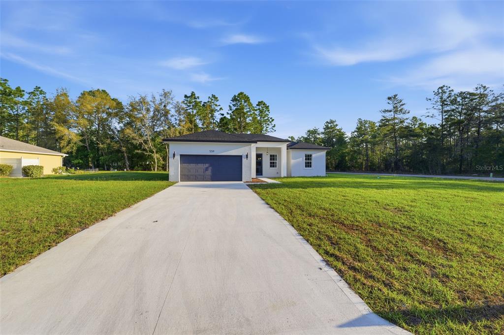 A single-story white house with a dark garage door sits on a grassy lot — 559 W HERONSBILL LANE, DUNNELLON, FL