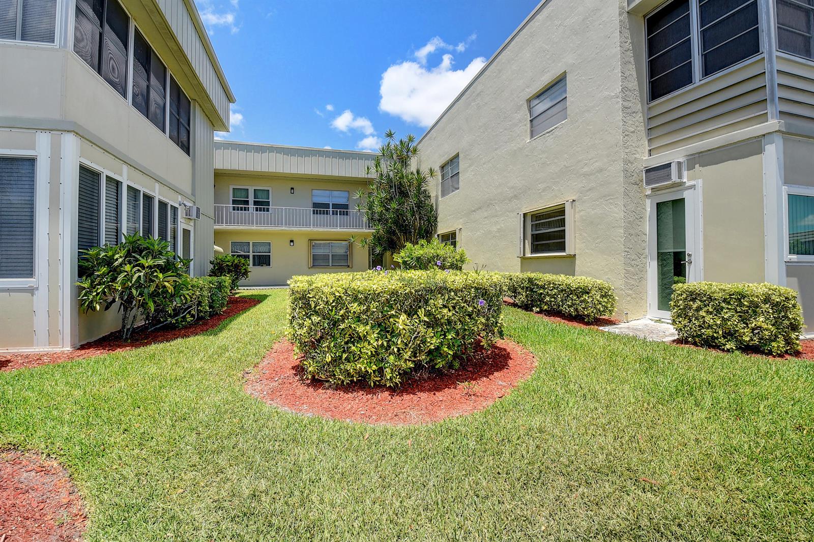 A grassy courtyard with manicured hedges sits between two multi-story apartment buildings under a sky — 318 Monaco Boulevard # G, Delray Beach FL 33446, DELRAY BEACH, FL