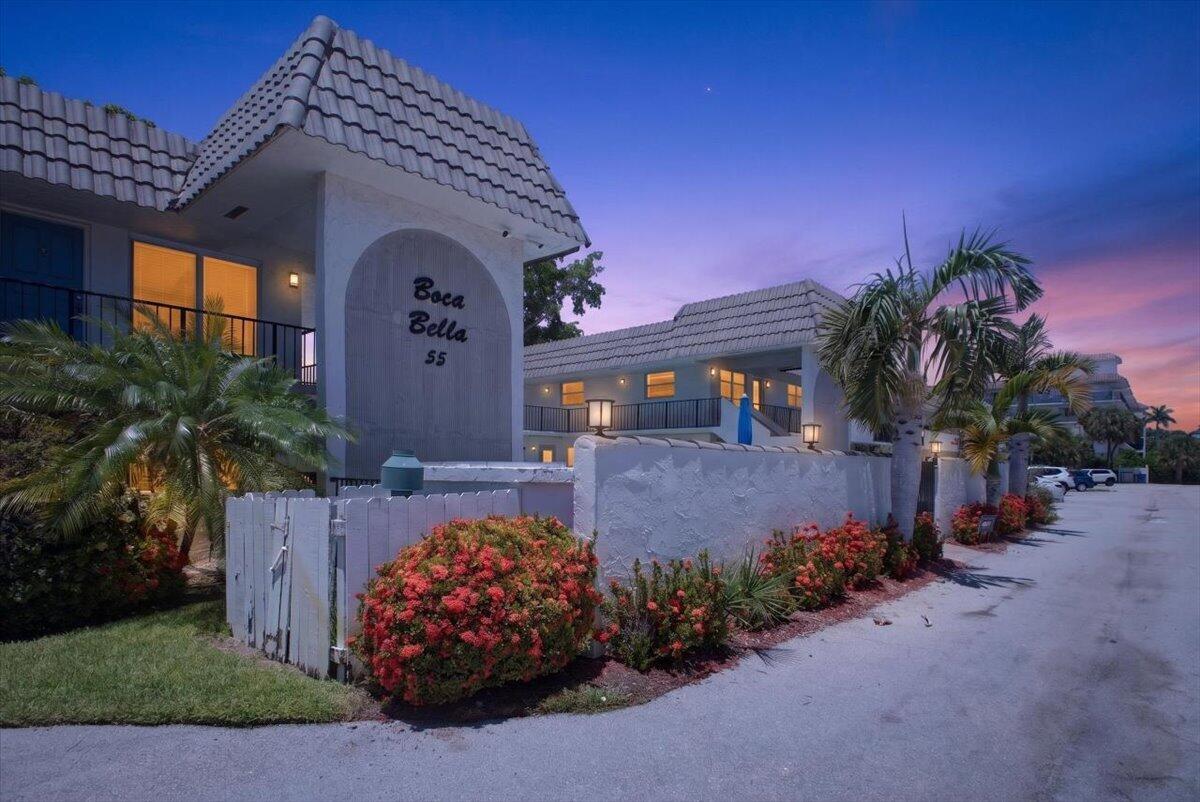Exterior view of a two-story apartment building with a white fence and flowering bushes — 55 SE 14th Street # 2060, Boca Raton FL 33432, BOCA RATON, FL