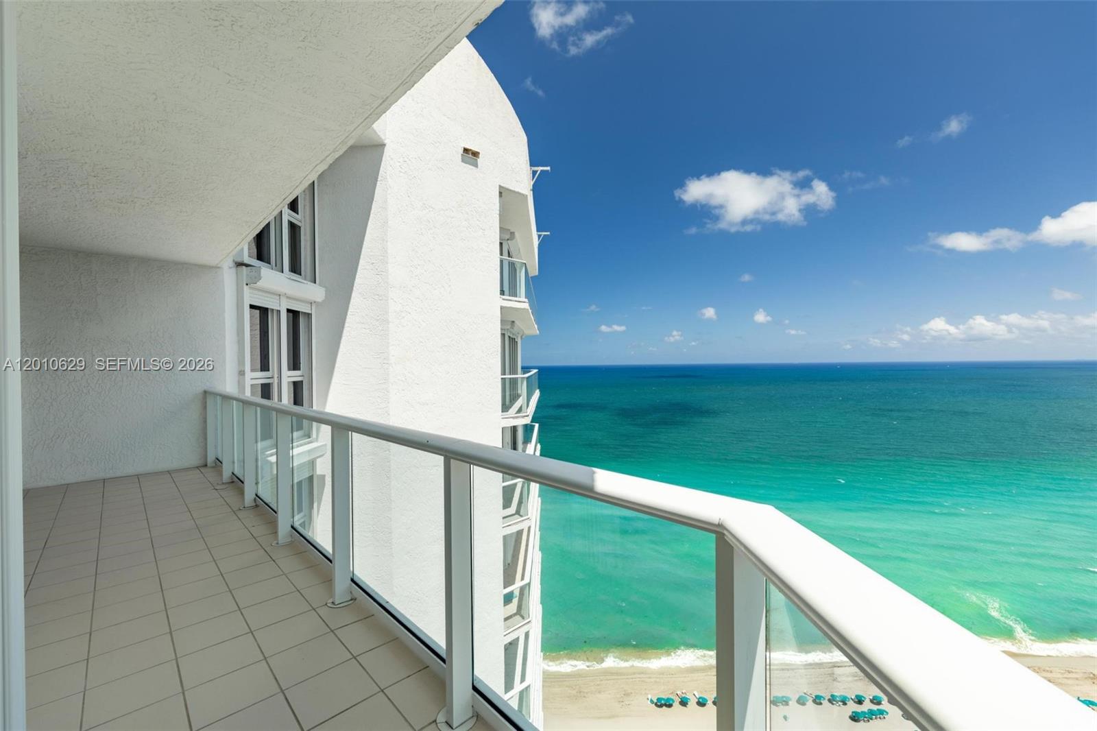 A high-rise balcony with a glass railing overlooks a sandy beach and the ocean — SUNNY ISLES BEACH, FL