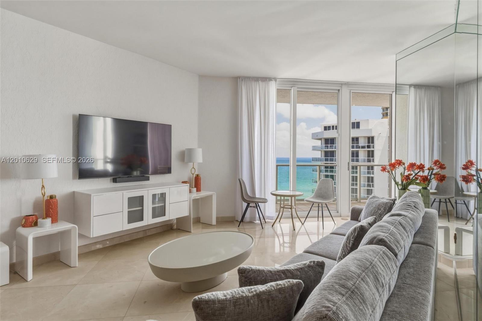 A living room features a grey sofa, white media console, and balcony with ocean views — SUNNY ISLES BEACH, FL