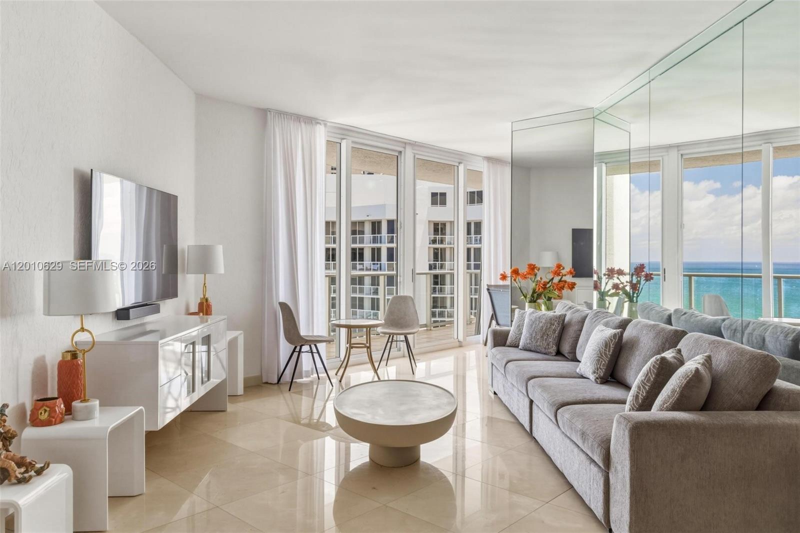 A bright living room features a gray sofa, white media console, and floor-to-ceiling windows — SUNNY ISLES BEACH, FL