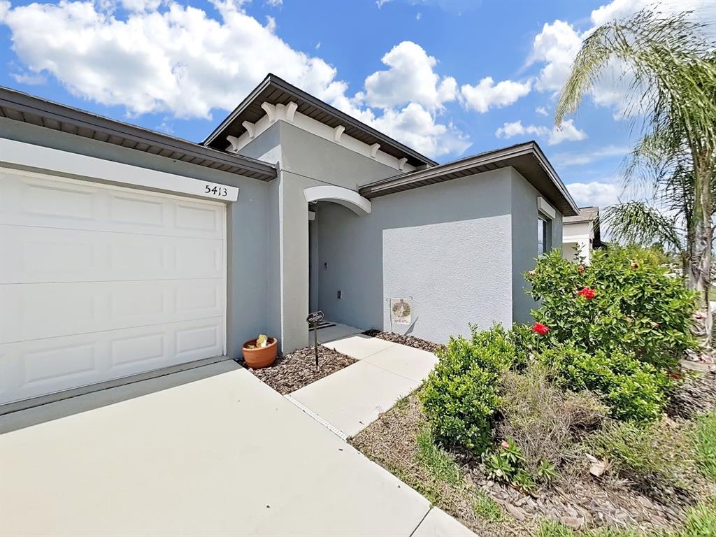 A single-story house exterior features a gray stucco facade, a white garage door, and landscaping — 5413 BLUE AZURE DRIVE, WIMAUMA, FL