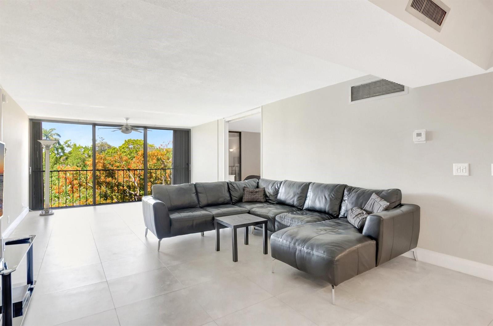 A living room with a large black sectional sofa, tile floors, and a balcony view — 501 SW 11th Place # 403, Boca Raton FL 33432, BOCA RATON, FL