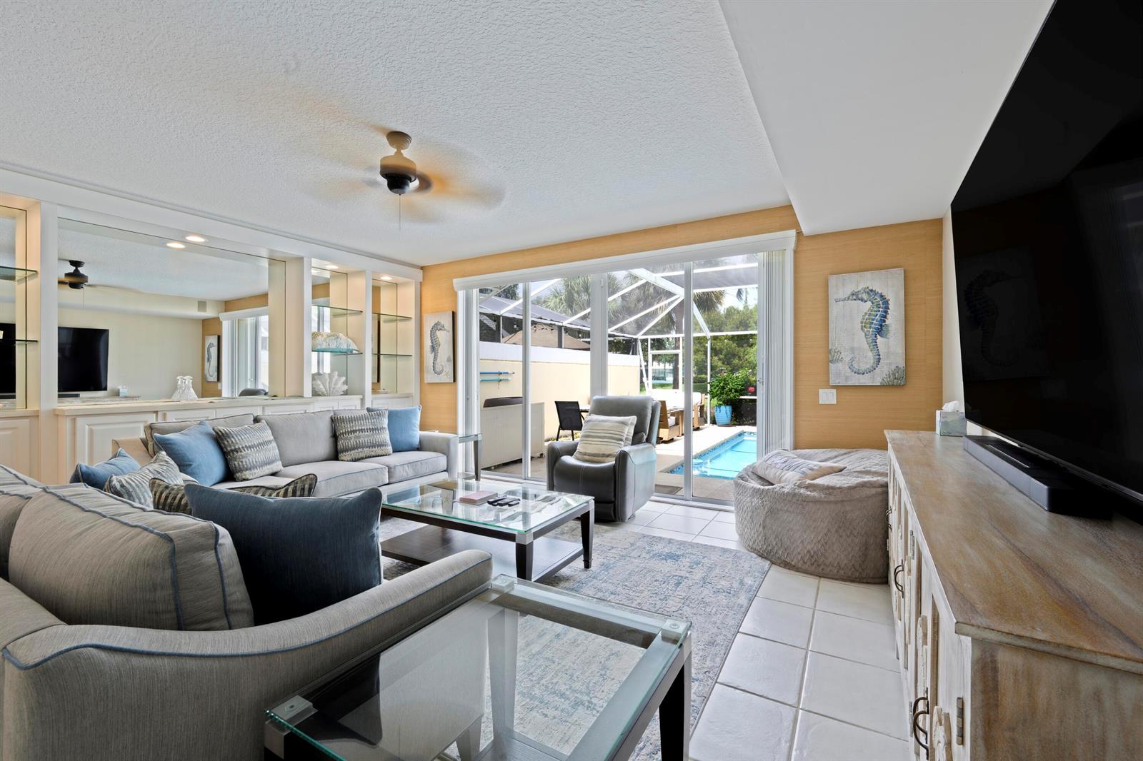 A living room features a grey sectional, glass coffee table, and sliding doors to a pool — 435 Capistrano Drive, Palm Beach Gardens FL 33410, PALM BEACH GARDENS, FL