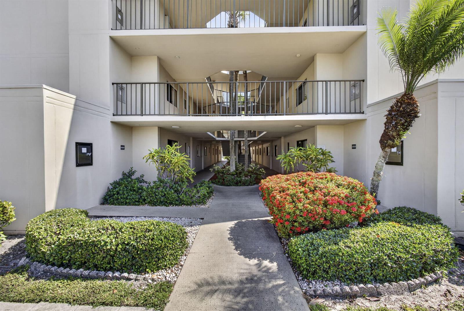 A concrete walkway leads to the open-air entrance of a multi-story apartment building with landscaping — 5340 Las Verdes Circle # 307, Delray Beach FL 33484, DELRAY BEACH, FL