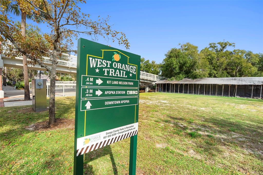 A green West Orange Trail sign stands in a grassy area near a pedestrian bridge — 215 E 5TH STREET, APOPKA, FL