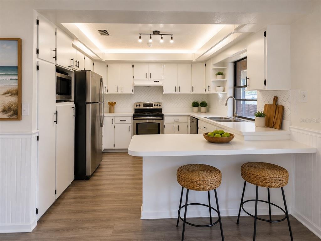 A white kitchen features stainless steel appliances, a breakfast bar with stools, and wood flooring — 3209 PAFKO DRIVE, SARASOTA, FL