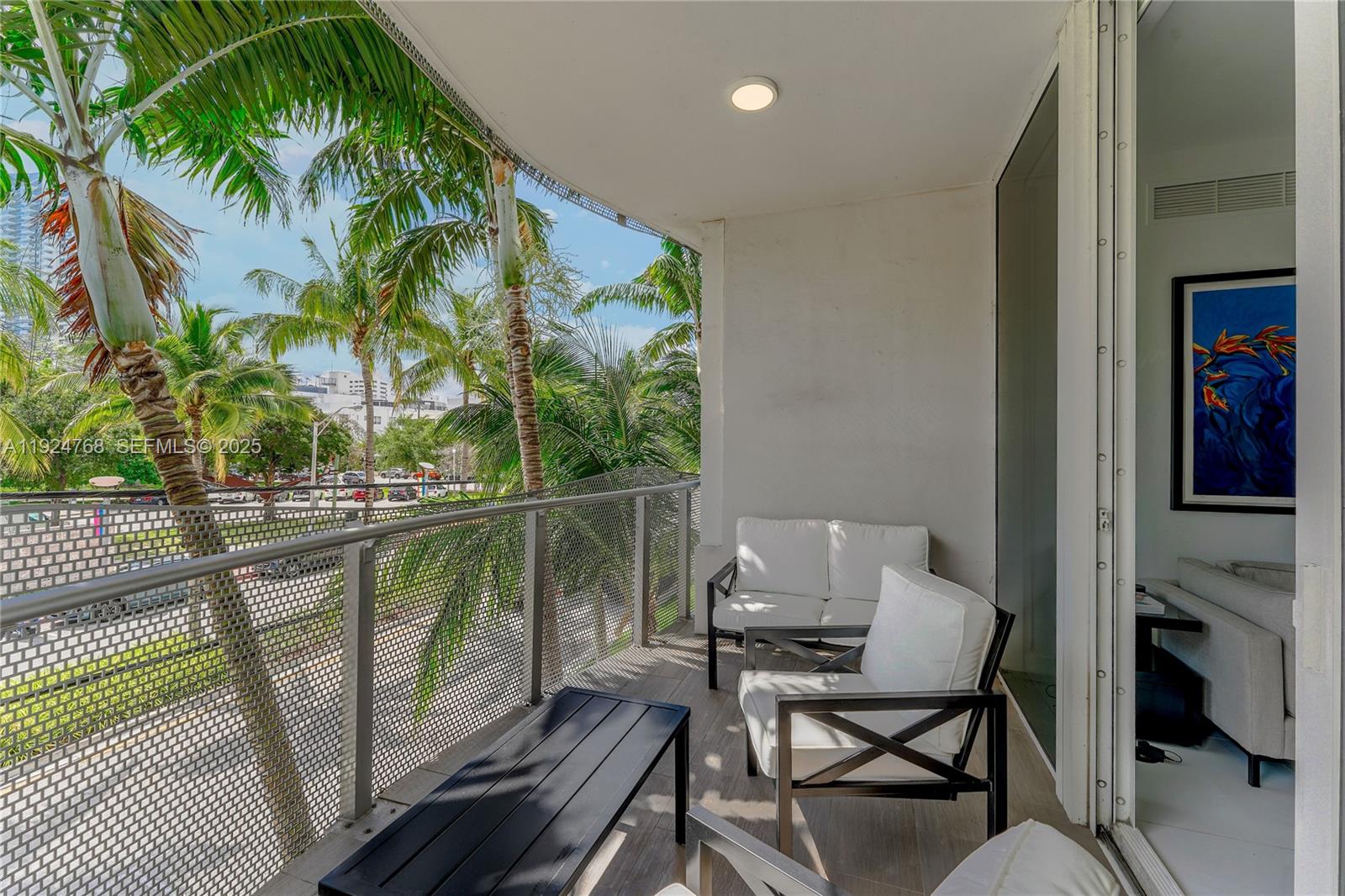 A balcony features white cushioned patio furniture, a black coffee table, and palm tree views — 2100 Park Ave # 201, Miami Beach FL 33139, MIAMI BEACH, FL