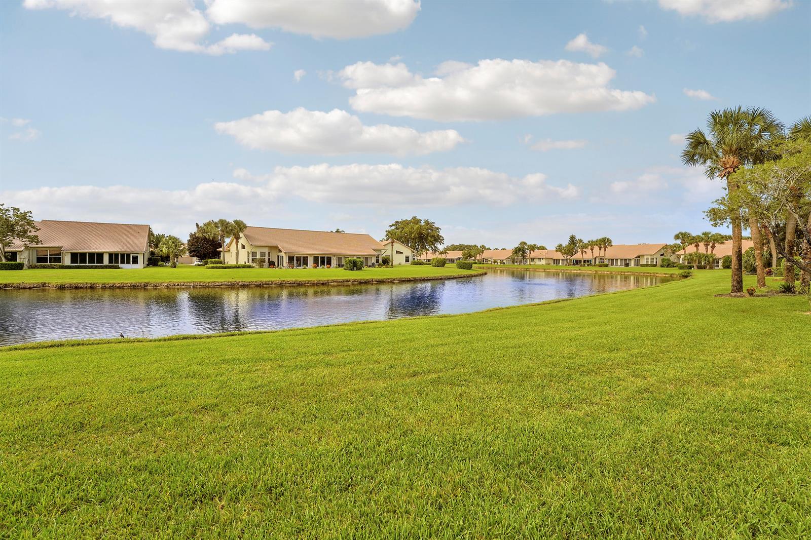 A grassy lawn borders a pond with residential houses in the background under a blue sky — 7136 Ashford Lane, Boynton Beach FL 33472, BOYNTON BEACH, FL