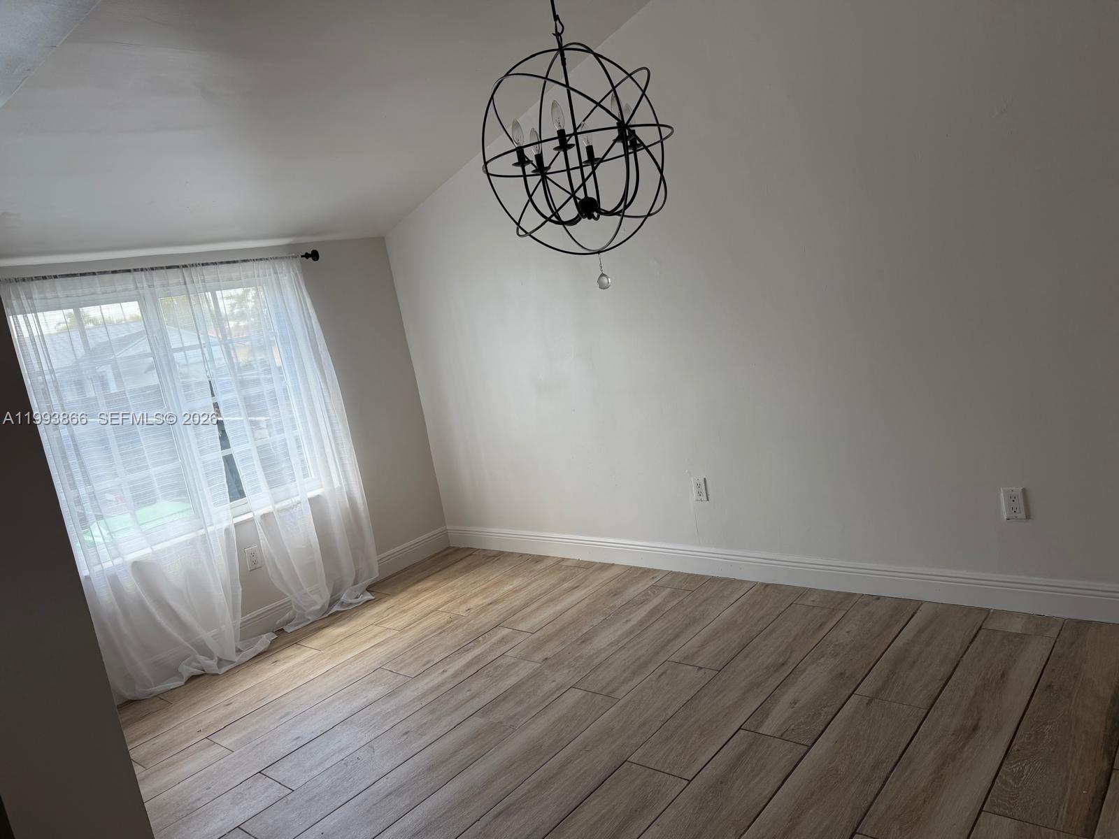 An empty room features light wood-look flooring, white walls, a large orb chandelier, and sheer curtains — 3427 SW 64th Ave, Miami FL 33155, MIAMI, FL