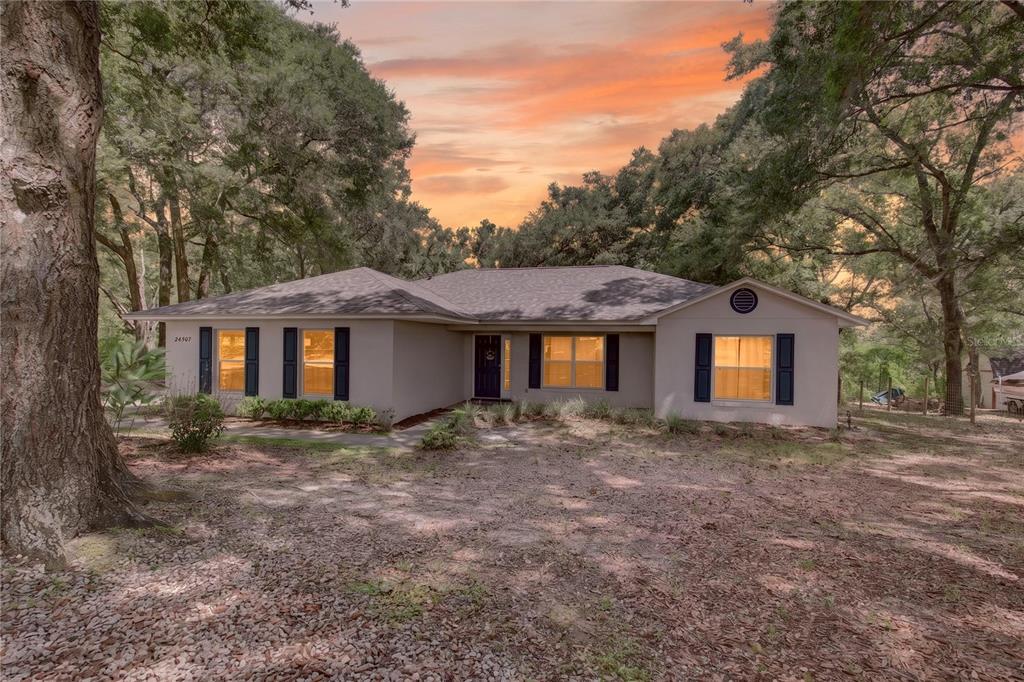 A single-story house with a light-colored exterior and dark shutters sits among large oak trees — 24307 ADAIR AVENUE, SORRENTO, FL