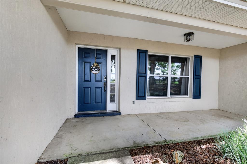 A covered concrete porch features a blue front door and a window with blue shutters — 24307 ADAIR AVENUE, SORRENTO, FL