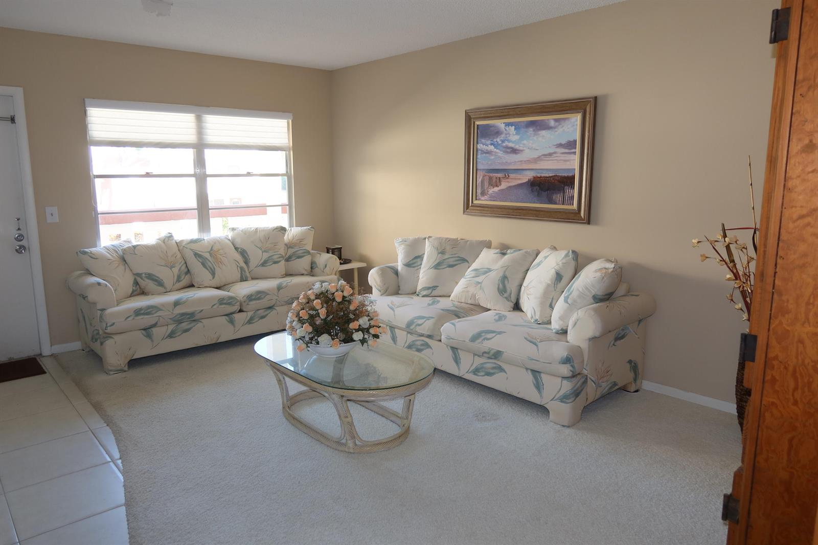 A living room features two floral sofas, a glass coffee table, and a beach-themed wall painting — 102 Brighton C, Boca Raton FL 33434, BOCA RATON, FL