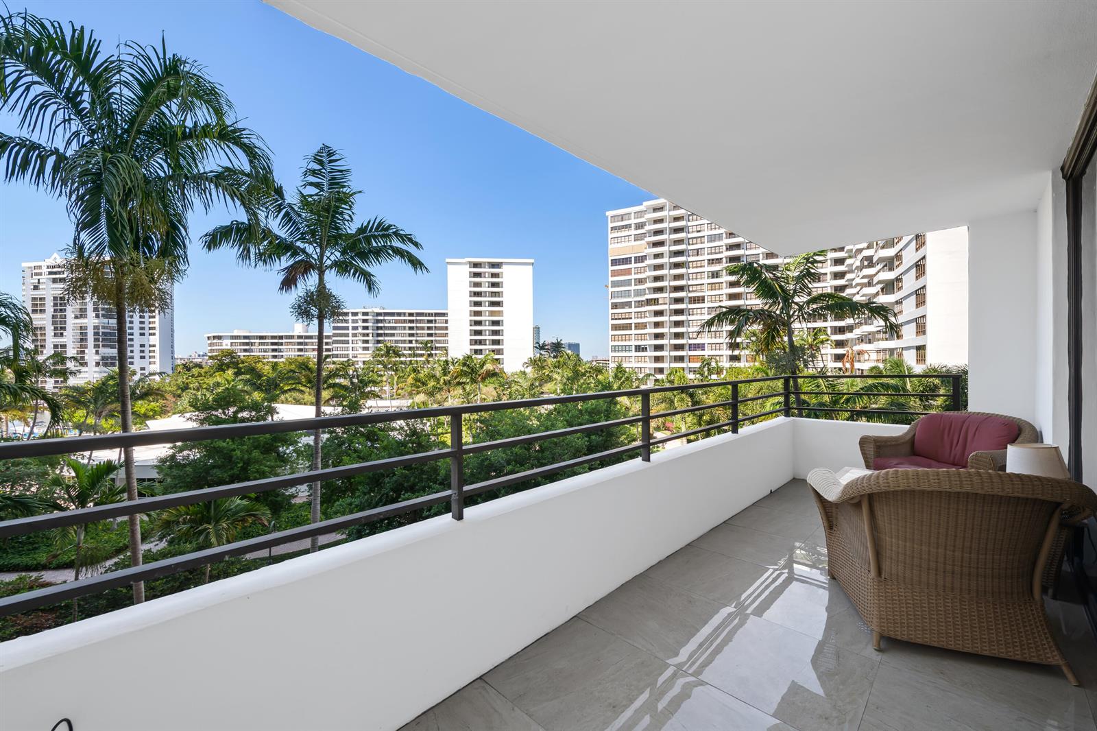 A balcony with tile flooring, wicker furniture, and a view of palm trees and buildings — 2500 Parkview Drive # 615, Hallandale Beach FL 33009, HALLANDALE BEACH, FL