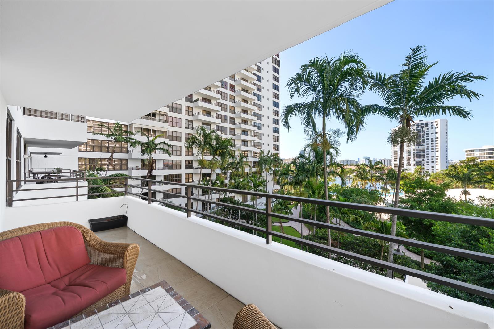 A balcony with wicker furniture overlooks a courtyard with palm trees and apartment buildings — 2500 Parkview Drive # 615, Hallandale Beach FL 33009, HALLANDALE BEACH, FL