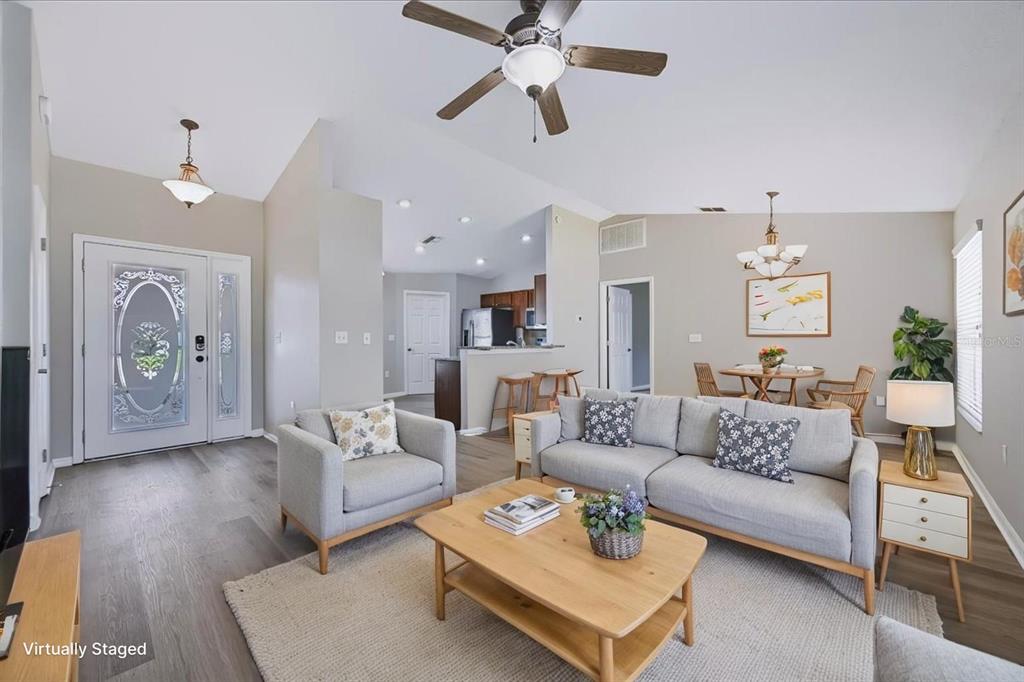 A living room with light wood floors, a grey sofa, and an open kitchen area — 8810 PEBBLEBROOKE DRIVE, LAKELAND, FL