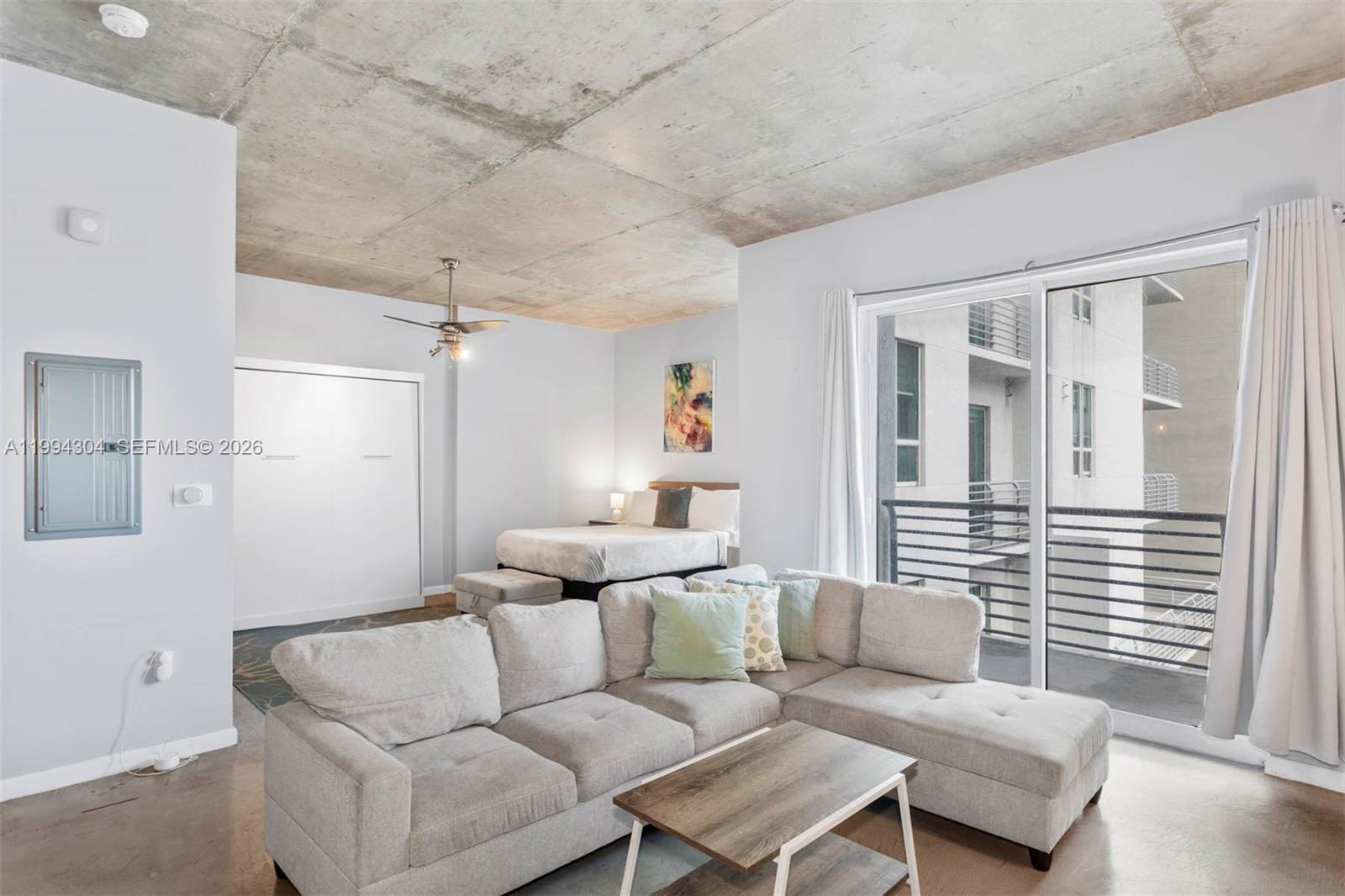 A studio apartment features a concrete ceiling, a sectional sofa, and a bed near a balcony — 234 NE 3rd St # 1106, Miami FL 33132, MIAMI, FL