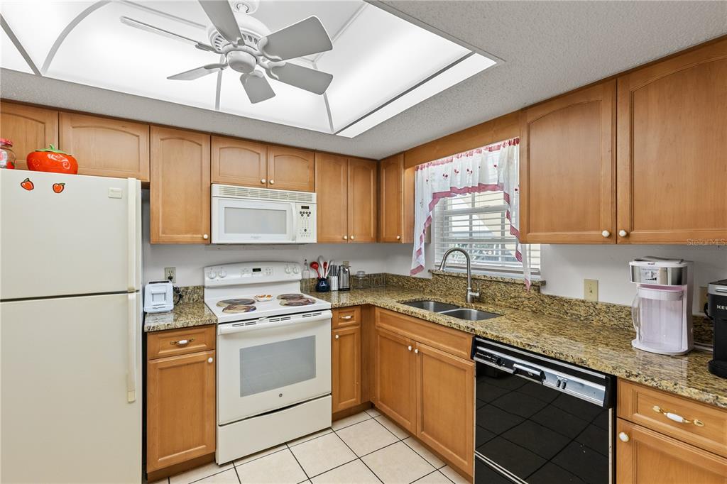 A kitchen features wood cabinets, granite countertops, white appliances, and a ceiling fan — 942 CAPRI ISLES BOULEVARD UNIT 105, VENICE, FL