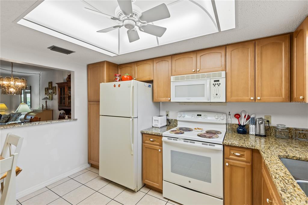 A kitchen features wood cabinets, granite countertops, white appliances, and a ceiling fan — 942 CAPRI ISLES BOULEVARD UNIT 105, VENICE, FL