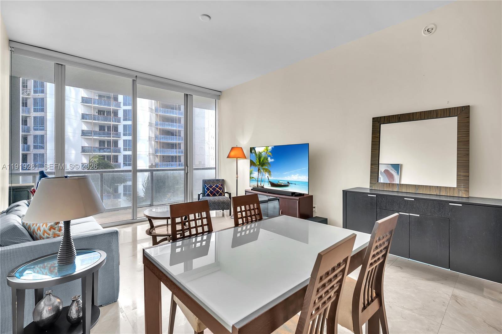 A living and dining area features a glass-topped table, sofa, television, and floor-to-ceiling windows — 6799 Collins Ave # 309, Miami Beach FL 33141, MIAMI BEACH, FL